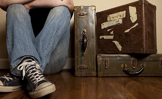 Floor level perspective of a young man wearing jeans, tennis shoes and a black tshirt sitting and leaning against a wall next to three old suitcases. Concept for travel or leaving for college.
